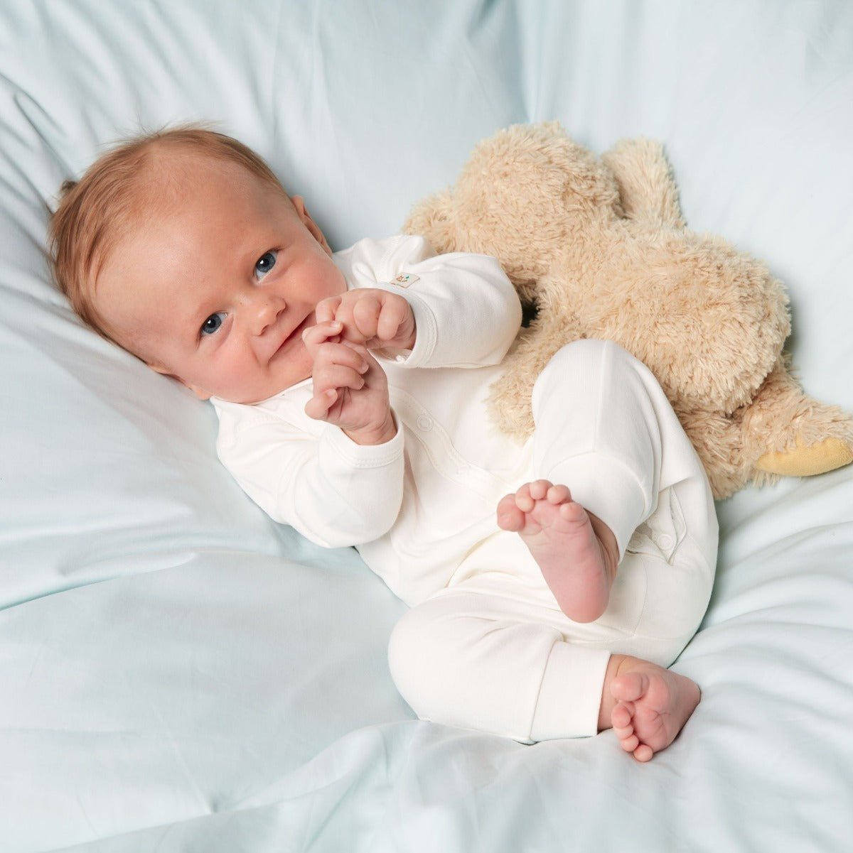 Bebis som ligger på ett täcke iklädd krämvit jumpsuit i ekologisk pimabomull