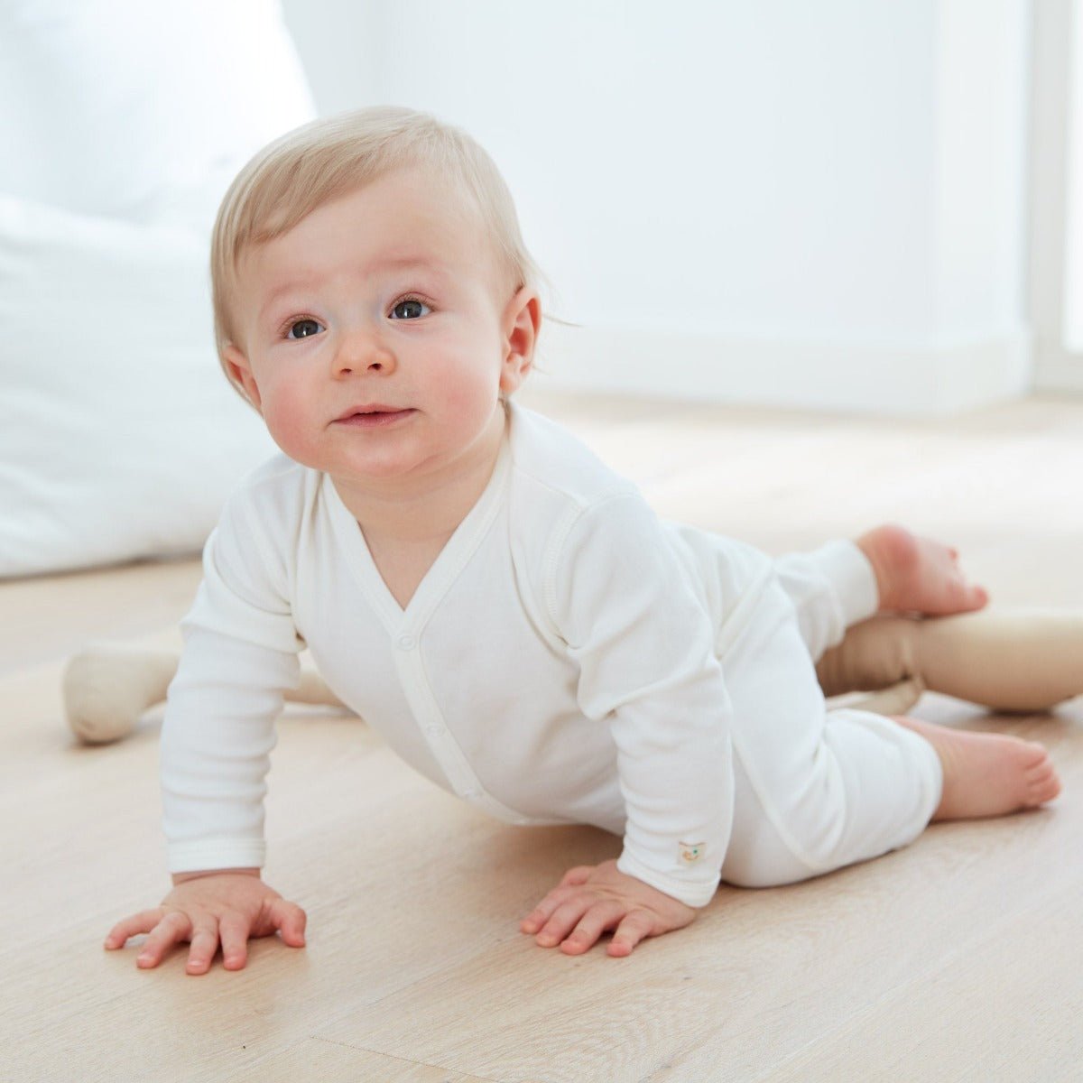 Bebis iklädd krämvit jumpsuit i ekologisk bomull som kryper på trägolv