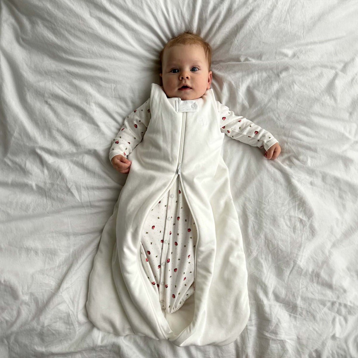 Bebis liggande på ett vitt täcke iklädd ekologisk sovpåse i krämvit bomull med dragkedja framtill