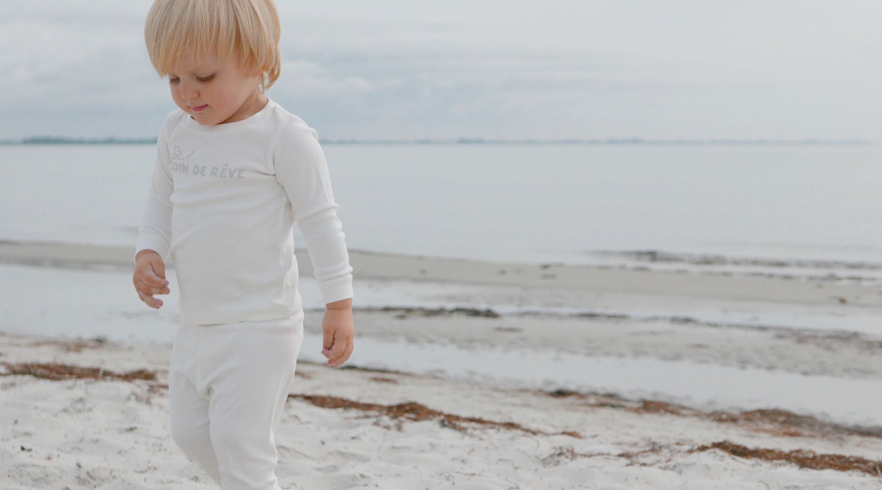 Söt pojke i tvådelad pyjamas går med bara fötter i sanden med havet i bakgrunden