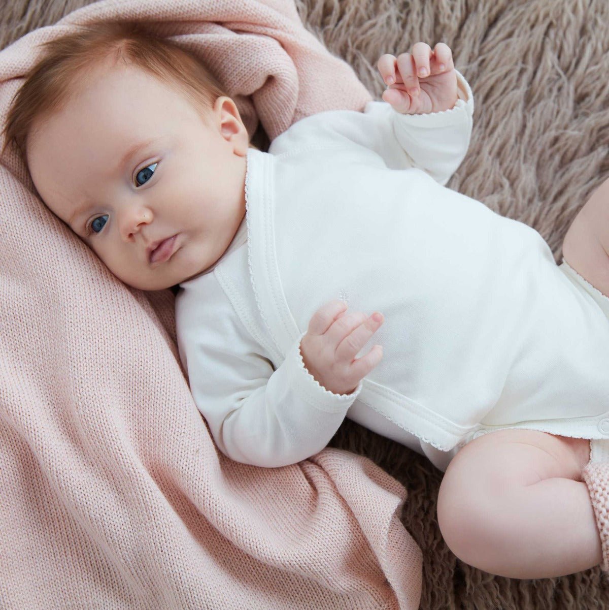 Liten bebis i vit ekologisk omlottbody som ligger på rosa stickad filt