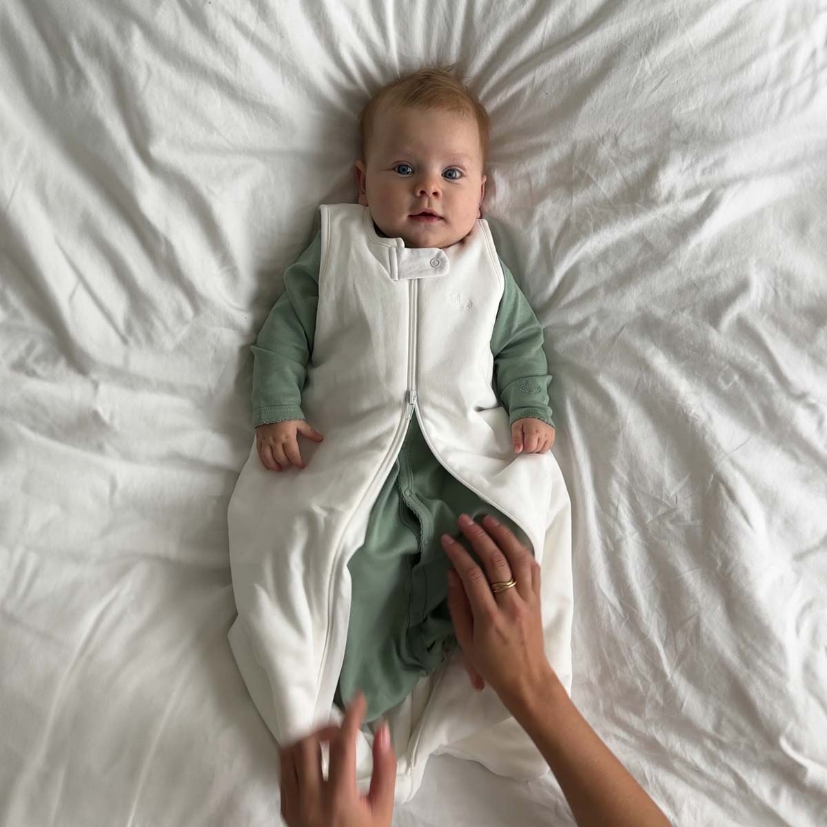 Bebis iklädd grön sovpåse (modell sleeping gown) och krämvit sovpåse (modell sleep sack) liggande på ett vitt täcke.