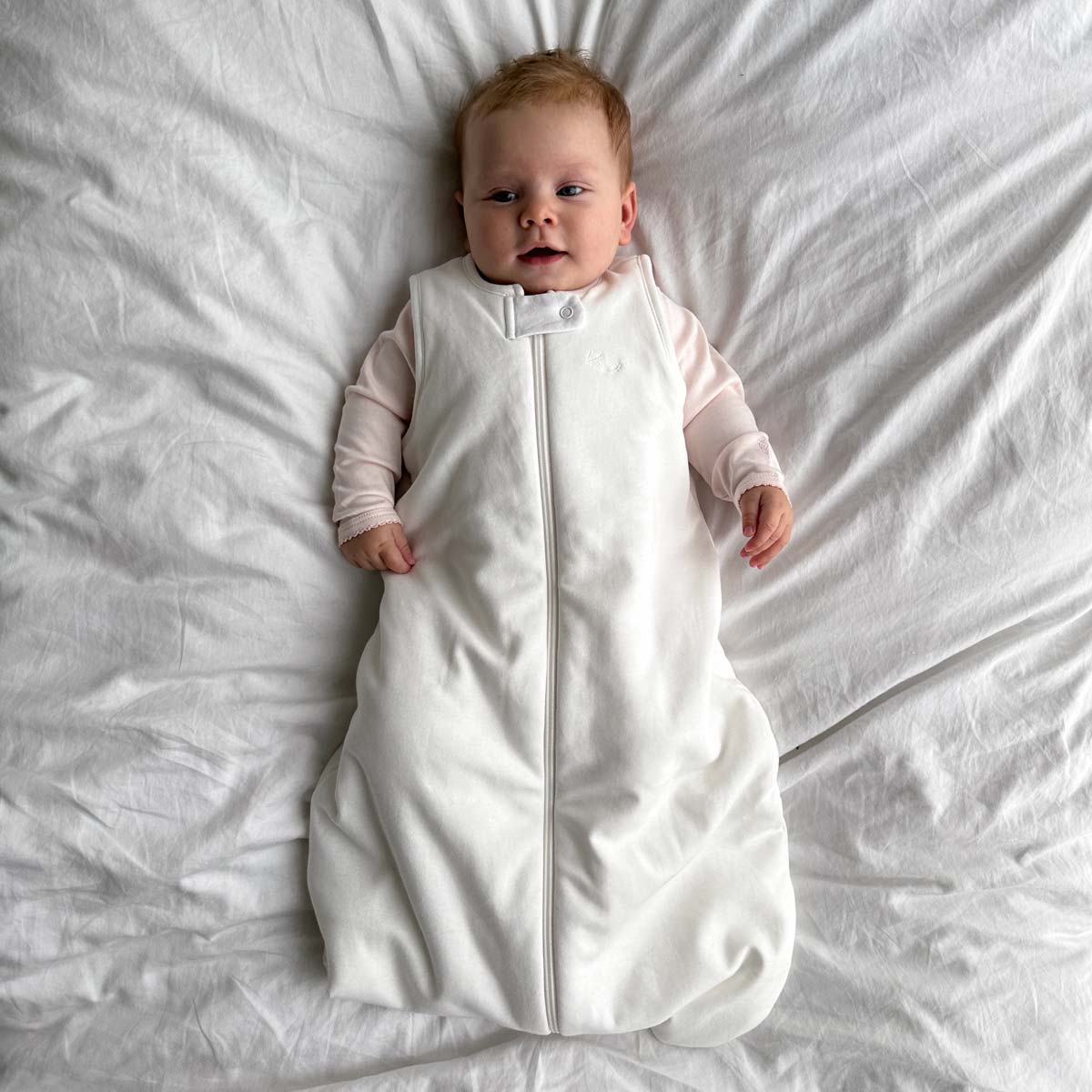 Bebis liggande på ett vitt täcke iklädd ekologisk sovpåse i krämvit bomull ovanpå rosa pyjamas eller sovpåse.