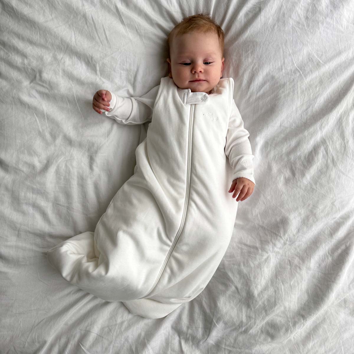 Bebis liggande på ett vitt täcke iklädd ekologisk sovpåse i krämvit bomull ovanpå pyjamas eller sovpåse o krämvit bomull.
